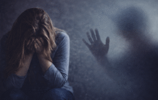 A distressed young woman sits with her head in her hands, wearing a grey shirt and jeans. A dark, blurred silhouette of a person with an outstretched hand appears on the right side of the image, creating a haunting and emotional atmosphere.
